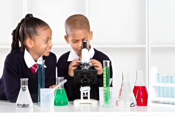 Young boy looking down a microscope, young girls looks on smiling. They are surrounded by lab glassware and chemicals